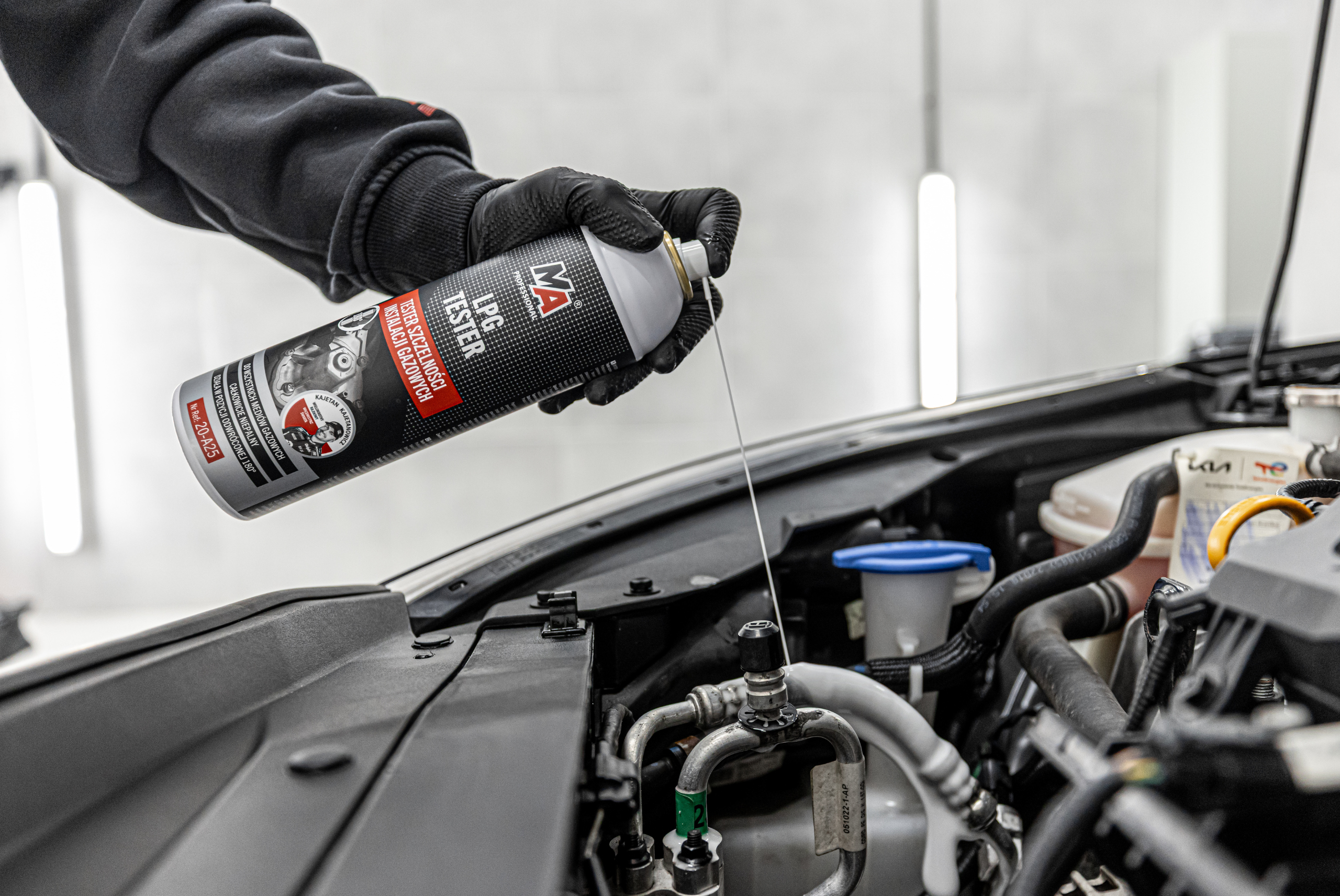 Osoba w czarnej rękawiczce aplikuje preparat MA Professional LPG Tester do układu klimatyzacji w komorze silnika samochodu. Aerozol jest rozpylany bezpośrednio na elementy instalacji w celu wykrywania nieszczelności.